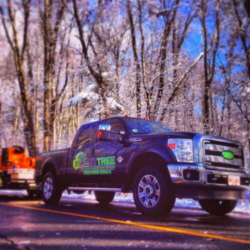 Kent Tree branded black pickup truck towing an orange stump grinder on a snow- and ice-coated tree-lined road.